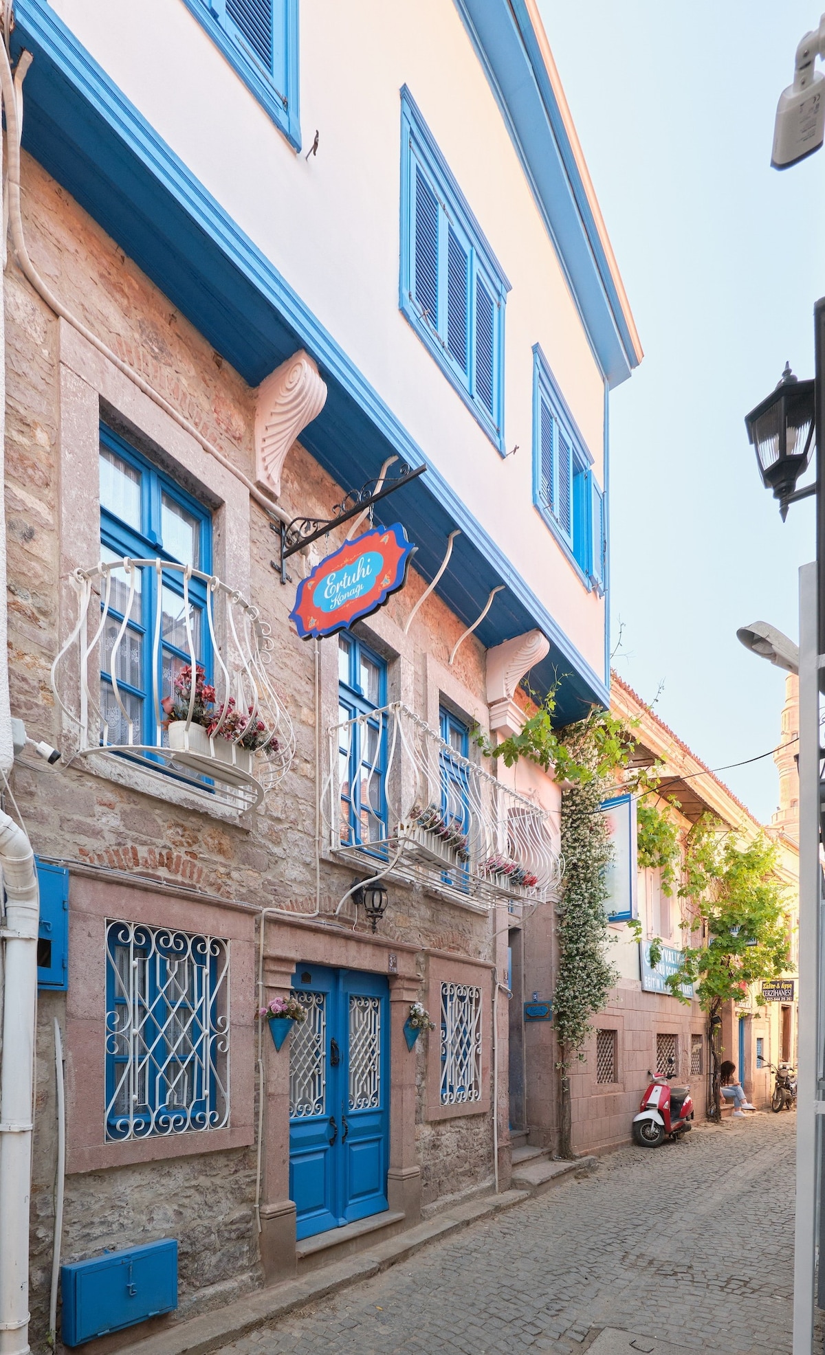 The historic stone house features a charming facade, highlighted by bright blue shutters and balcony railings adorned with potted flowers. The narrow cobblestone street contributes to the quaint atmosphere, while a red scooter is visible nearby, adding a touch of local life.