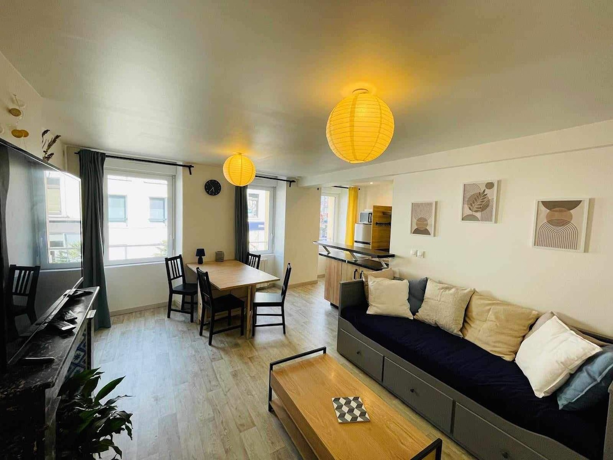 A bright living area is showcased with a dining table set for four and a sofa adorned with cushions. Two paper lanterns provide soft lighting, while windows enhance natural light. An open kitchen area is visible in the background, contributing to a spacious feel.