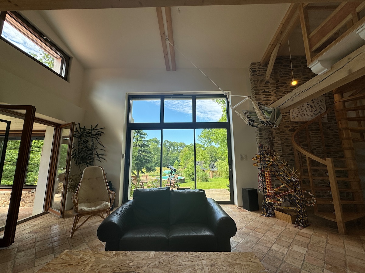 A spacious living area is illuminated by large windows, offering views of the lush greenery outside. A hammock is visible to one side, and a staircase leads to an upper level. Comfortable seating is arranged in the center, complemented by a rustic wooden coffee table.
