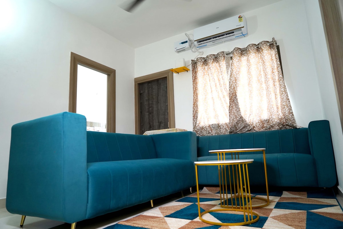 A tastefully decorated living area features two teal sofas and a gold-finished coffee table on a geometric-patterned rug. Natural light enters through large windows adorned with sheer curtains, and an air conditioning unit is mounted on the wall for added comfort.