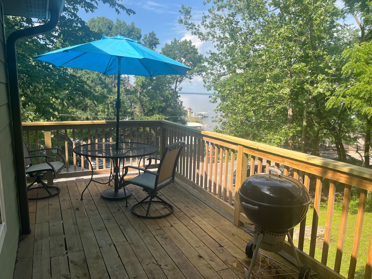A wooden deck features a round table surrounded by four chairs, an umbrella providing shade in a vibrant blue color, and a grill positioned nearby. Lush trees create a sense of privacy, with a view of the serene surroundings beyond.
