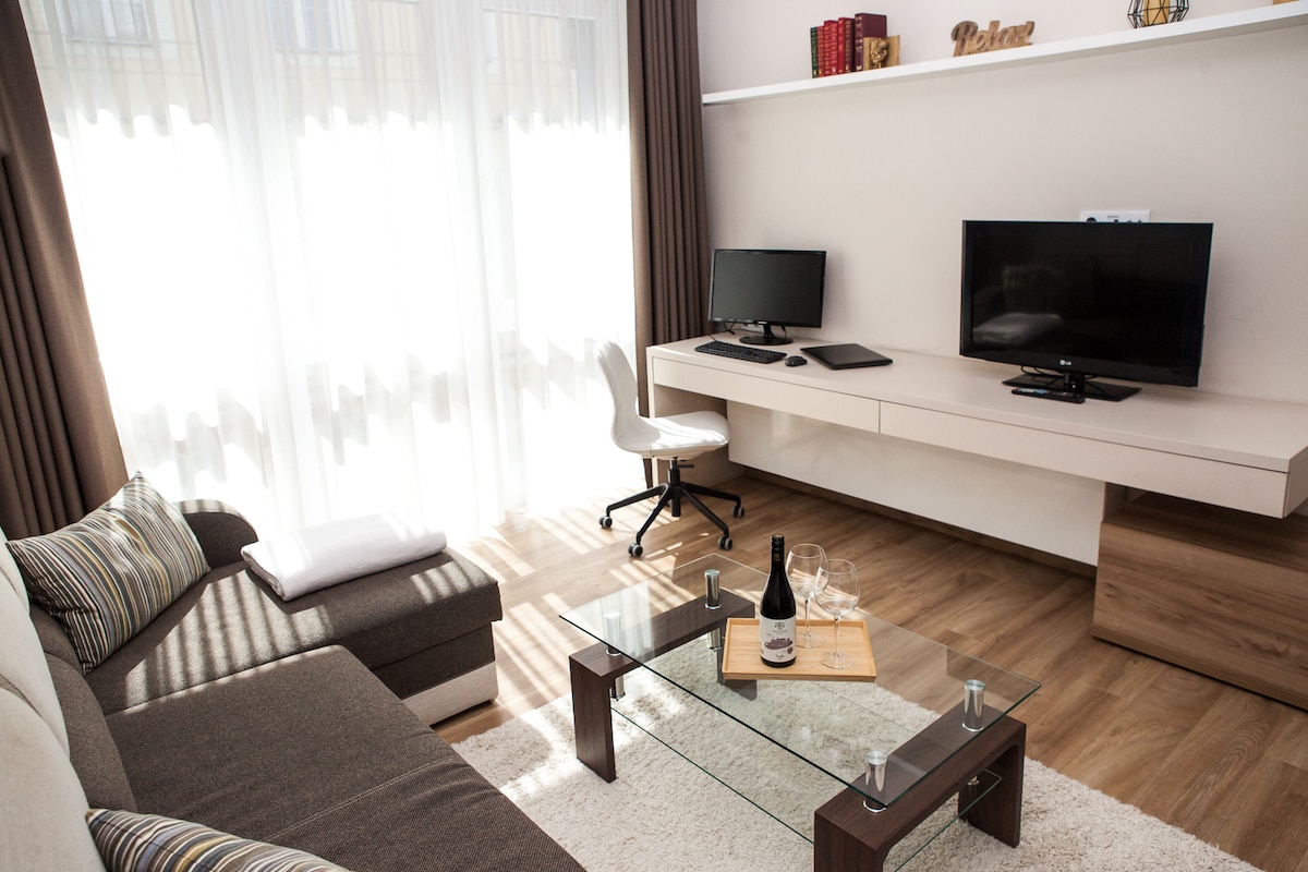 A light-filled living area showcases a comfortable sectional sofa paired with a glass coffee table. Two monitors are positioned on a mounted desk against the wall. Dark curtains frame the large window, creating a cozy and functional space for relaxation or work.
