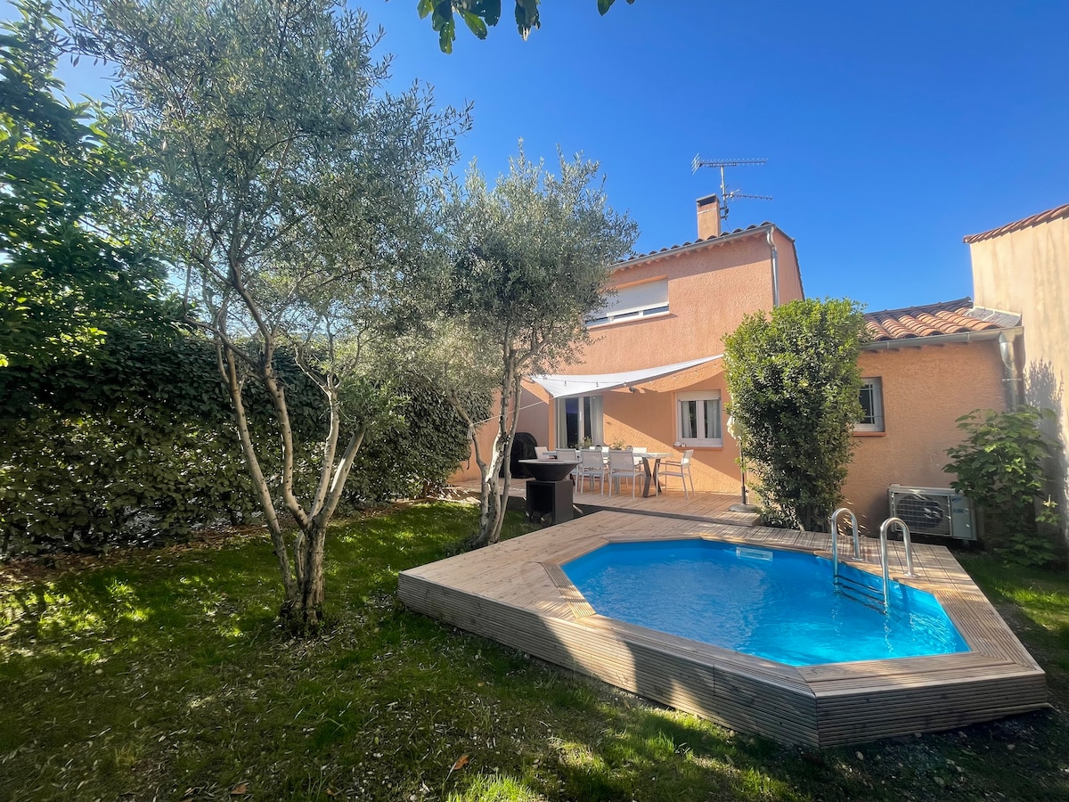 A charming house is depicted surrounded by lush greenery, featuring a small pool with a wooden deck. Olive trees provide shade, and a dining area is visible under a retractable awning. Clear blue skies enhance the inviting atmosphere of the outdoor space.
