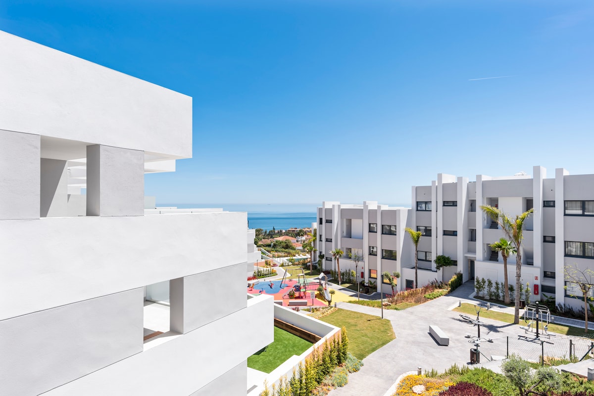 The image captures a modern residential area featuring white buildings under a clear blue sky. A view of the sea is visible in the background, complemented by landscaped gardens and colorful flowerbeds. Outdoor recreational spaces, including a pool, can be seen in the well-maintained grounds.