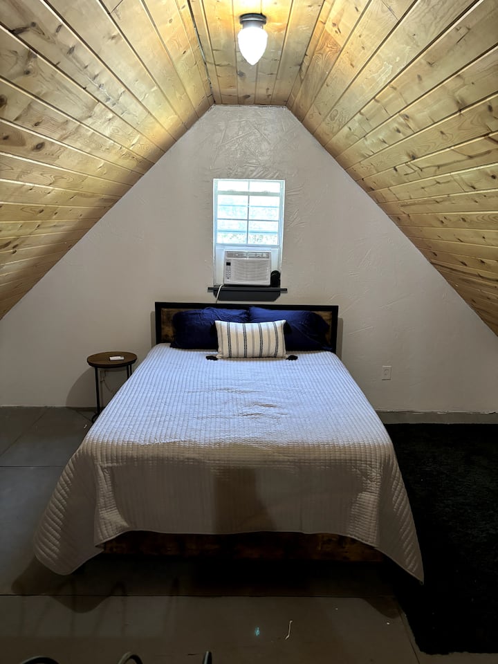 Manželská posteľ s jedným odkladacím stolíkom.  Okenná  klimatizačná jednotka pre podkrovie. 