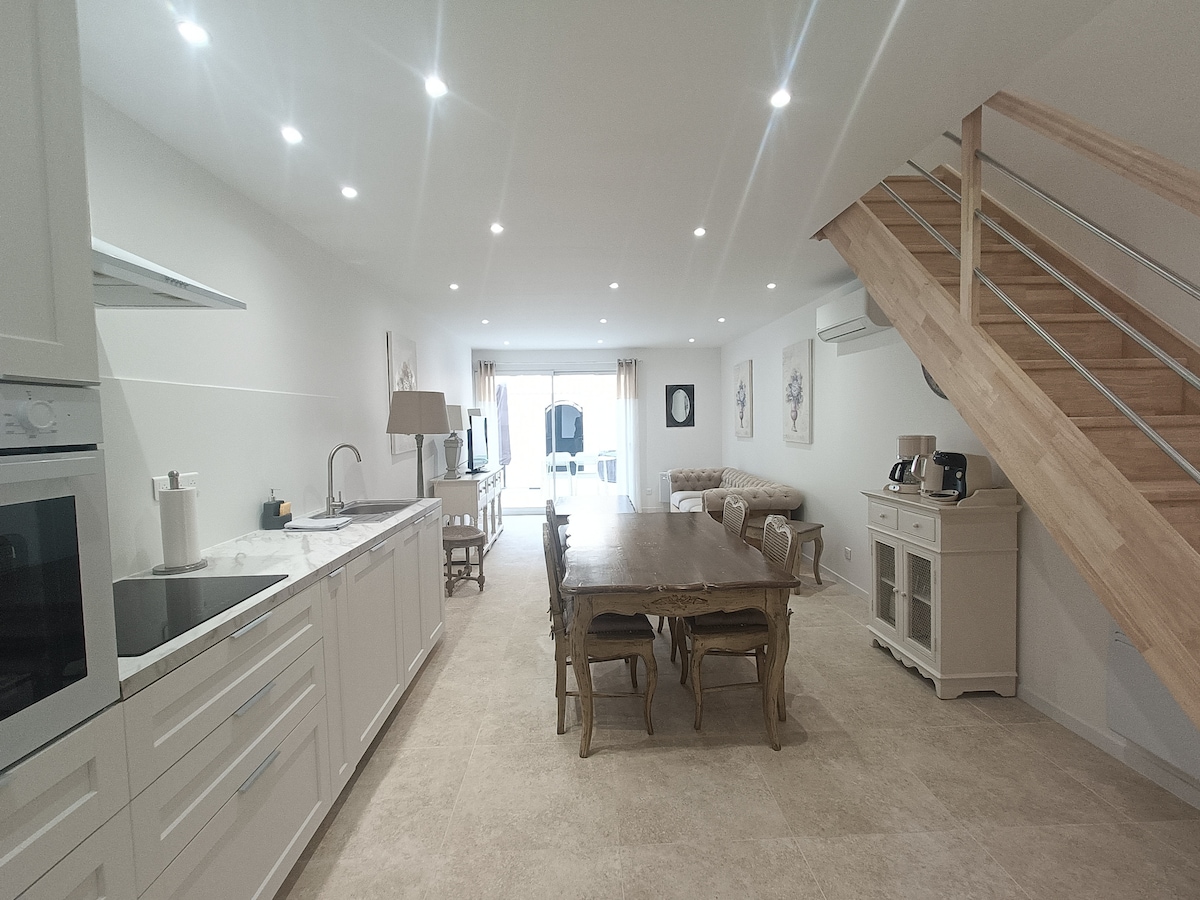 A spacious kitchen and dining area features a long wooden table surrounded by chairs. Modern kitchen appliances are integrated into white cabinetry, complemented by a light countertop. Natural light fills the room through large glass doors, which lead to the outdoor space, enhancing the inviting atmosphere.