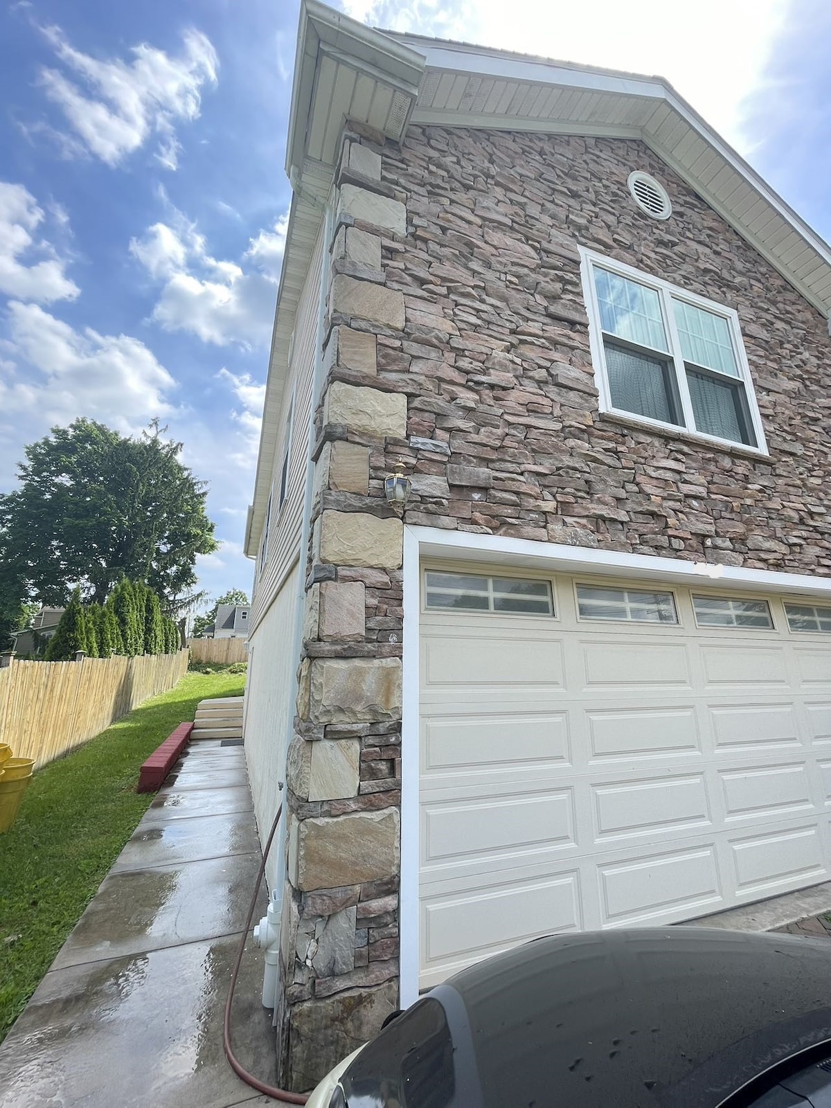 The side of a two-story home is depicted, showcasing a combination of stone and siding materials. Large windows are present, allowing natural light to enter. A smooth driveway extends alongside the house, leading to a standard garage door, which adds functionality to the exterior.