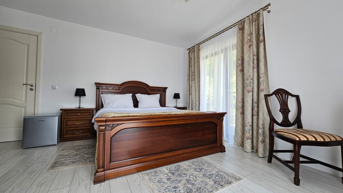 A well-furnished bedroom features a wooden bed with a light-colored duvet, flanked by two nightstands with lamps. A chair with a striped cushion complements the space, while a small refrigerator is positioned next to a balcony door. Natural light streams through the sheer curtains.