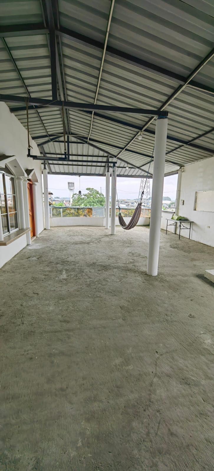 An open outdoor space features a concrete floor and a covered roof, supported by white pillars. A hammock hangs from the ceiling, and large windows allow natural light to fill the area. Minimal furnishings are present, ensuring a spacious feel.