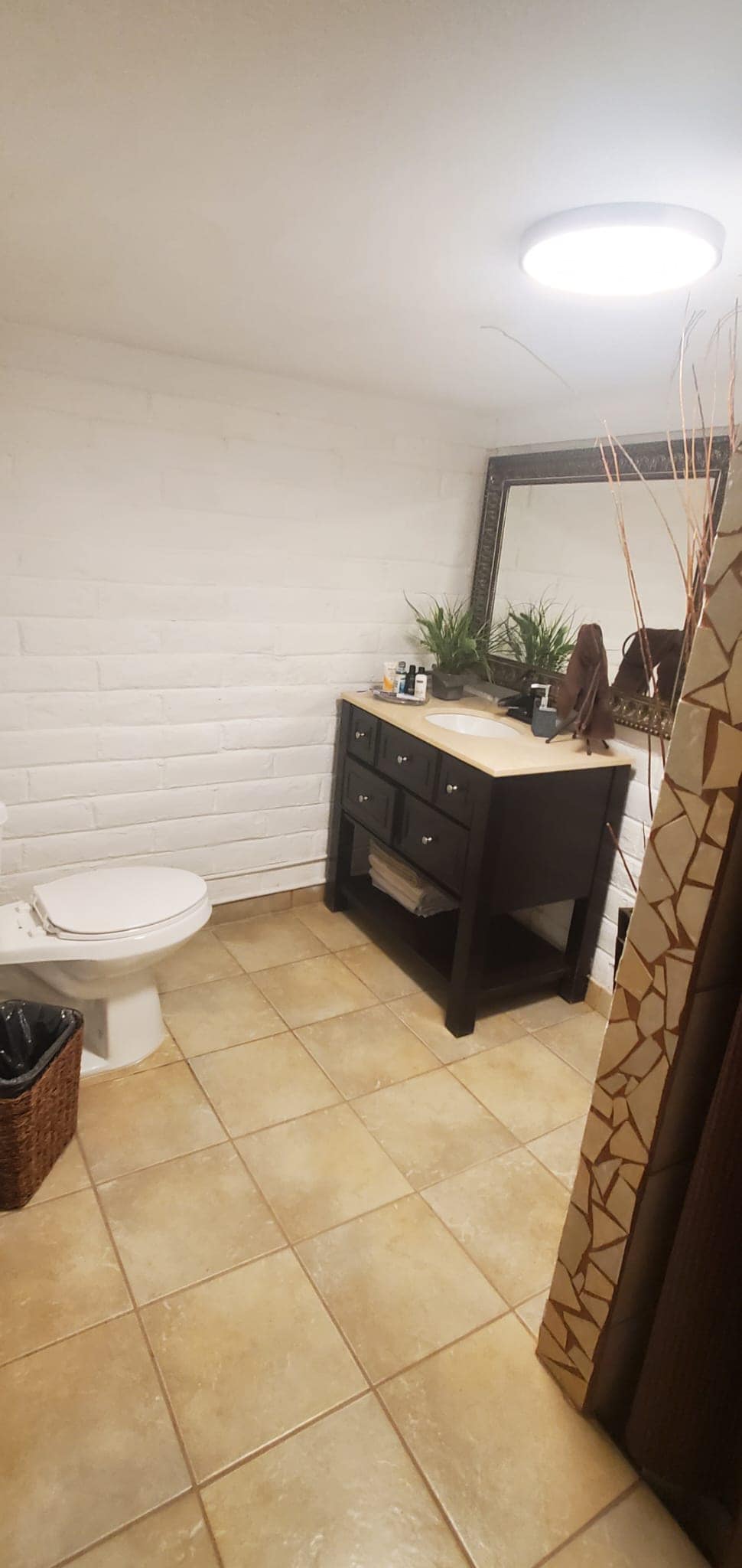 A well-appointed bathroom features light-colored brick walls and elegant tile flooring. A dark wooden vanity with storage drawers is positioned under a large mirror, complemented by decorative plants. A waste bin is visible, along with a shower curtain that adds texture to the space.