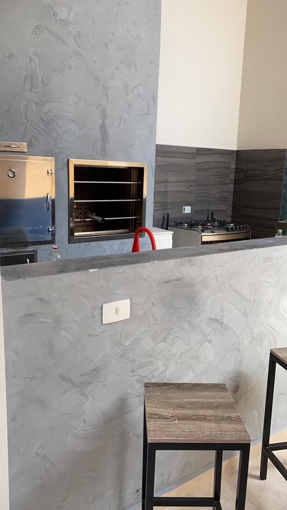 A modern kitchen space is presented featuring sleek gray walls and a polished countertop. The kitchen includes a gas stove and an oven set within dark cabinetry. In the foreground, two wooden bar stools are positioned near the countertop, providing a casual dining area.