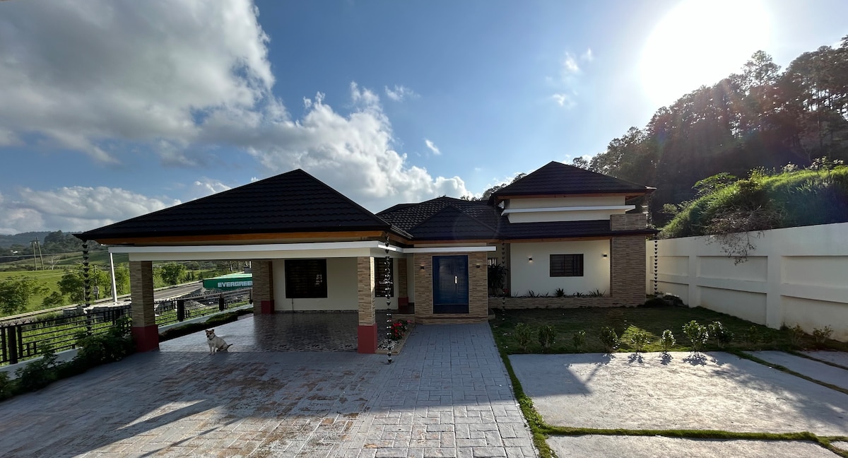 The exterior of the house features a contemporary design with a dark roof and light-colored walls. A spacious driveway is visible, bordered by manicured landscaping. Nearby hills can be seen in the background, contributing to the tranquil setting.
