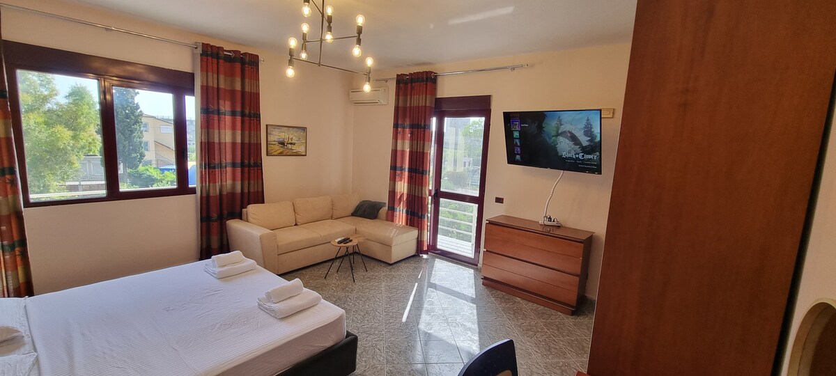 A bright and airy bedroom features a spacious bed and a comfortable sofa, positioned near large windows that provide natural light. Decorative curtains add a touch of color, while a modern light fixture hangs from the ceiling. A flat-screen TV is mounted on the wall, enhancing the room's functionality.