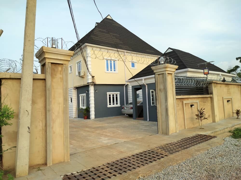 A stylish two-story building is displayed behind a secure, gated entrance. Barbed wire lines the top of the wall, while a smooth driveway leads to the car parked near the entrance. The exterior features a blend of light and dark colors, contributing to its modern appearance.