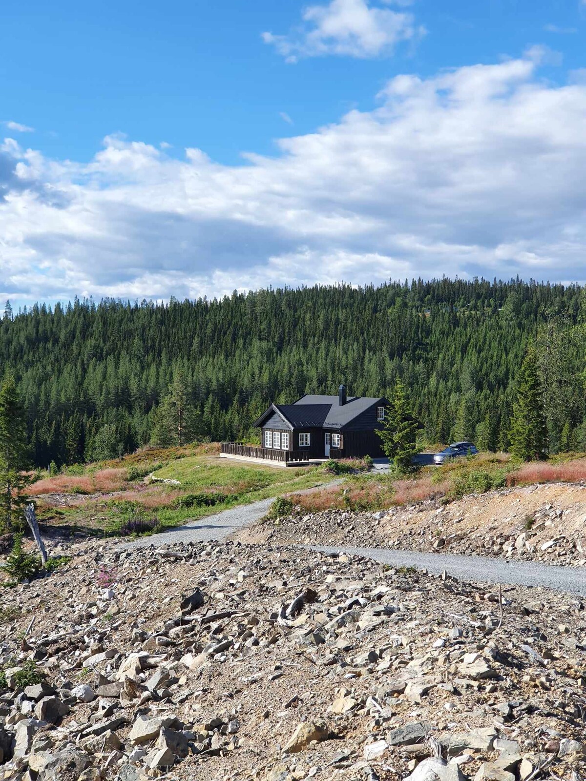 A black cabin is situated on a hillside, surrounded by lush green forests. A gravel path leads up to the cabin, framed by rocky terrain and vibrant vegetation. The sky is partly cloudy, with patches of blue visible amongst the clouds.