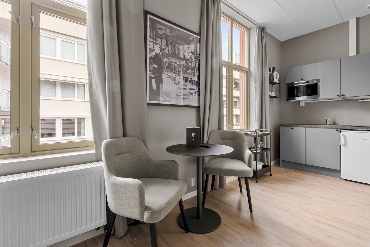 A well-lit dining area is presented, featuring a circular table with two chairs. Large windows allow natural light to fill the space, while subtle artwork adorns the wall. A modern kitchen is visible in the background, equipped with basic appliances.