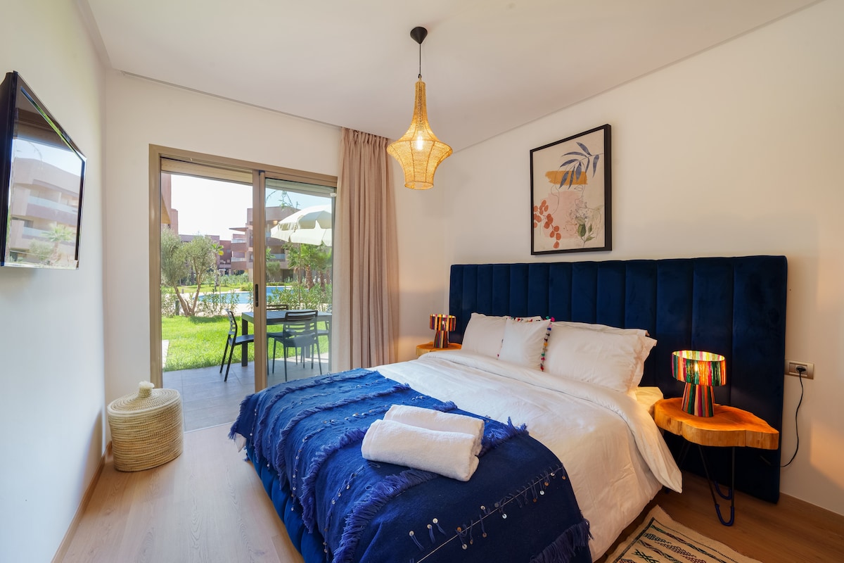 A spacious bedroom features a large bed with a blue velvet headboard and crisp white linens. A green garden and pool can be seen through sliding glass doors. A pendant light hangs above, while a small seating area with a table and chairs enhances the inviting atmosphere.