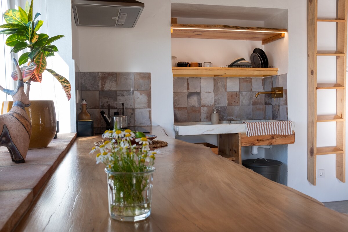 A well-appointed kitchen features wooden countertops with a central island. Natural light filters through a nearby window, illuminating the textured stone backsplash. Shelves hold dishware, while a small flower arrangement adds a touch of greenery to the inviting space.