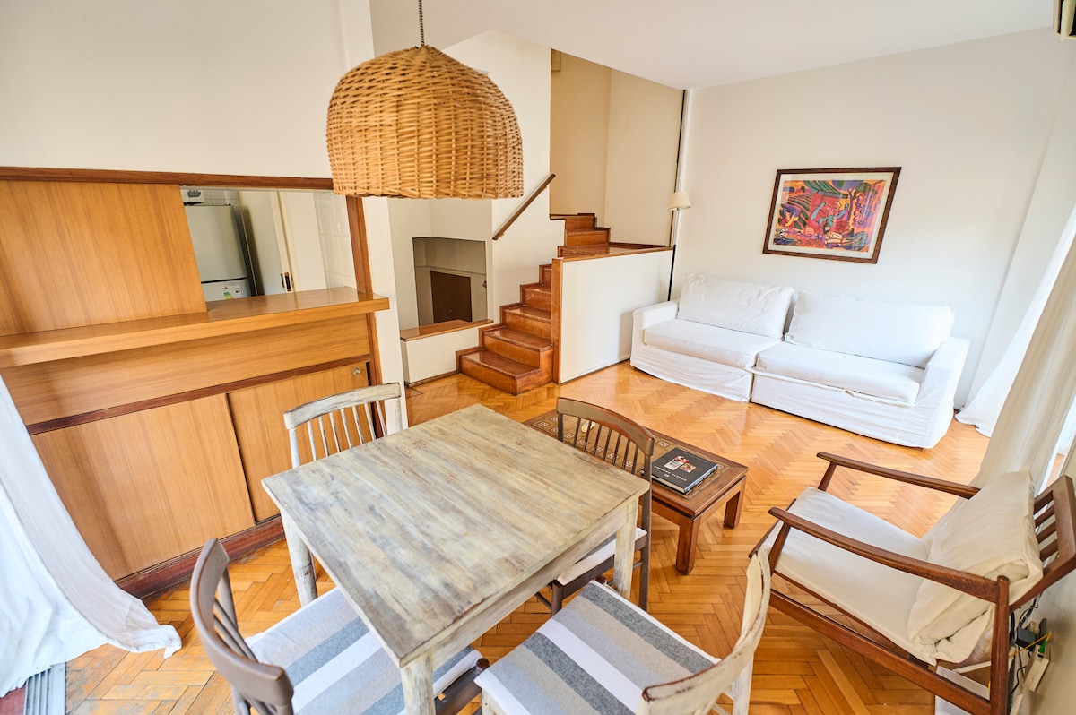 A spacious living area showcases wooden flooring and a cozy seating arrangement with a white sofa and wooden chairs. A rustic dining table is positioned beside a staircase leading to the upper level, and natural light enters through large windows, enhancing the open feel.