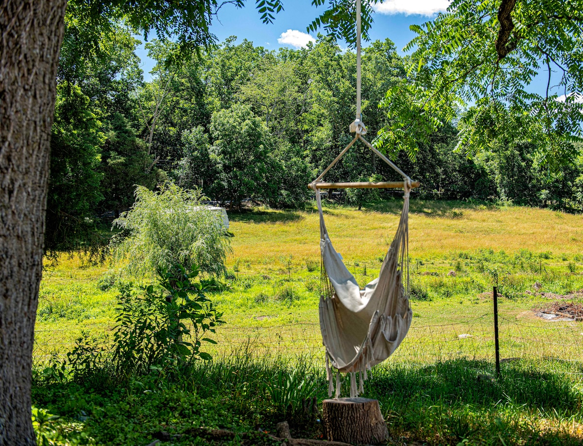 A cozy hammock is suspended from a sturdy frame, nestled among vibrant green grass and surrounded by lush trees. A serene field with tall grass stretches in the background, offering a peaceful view of the natural landscape.
