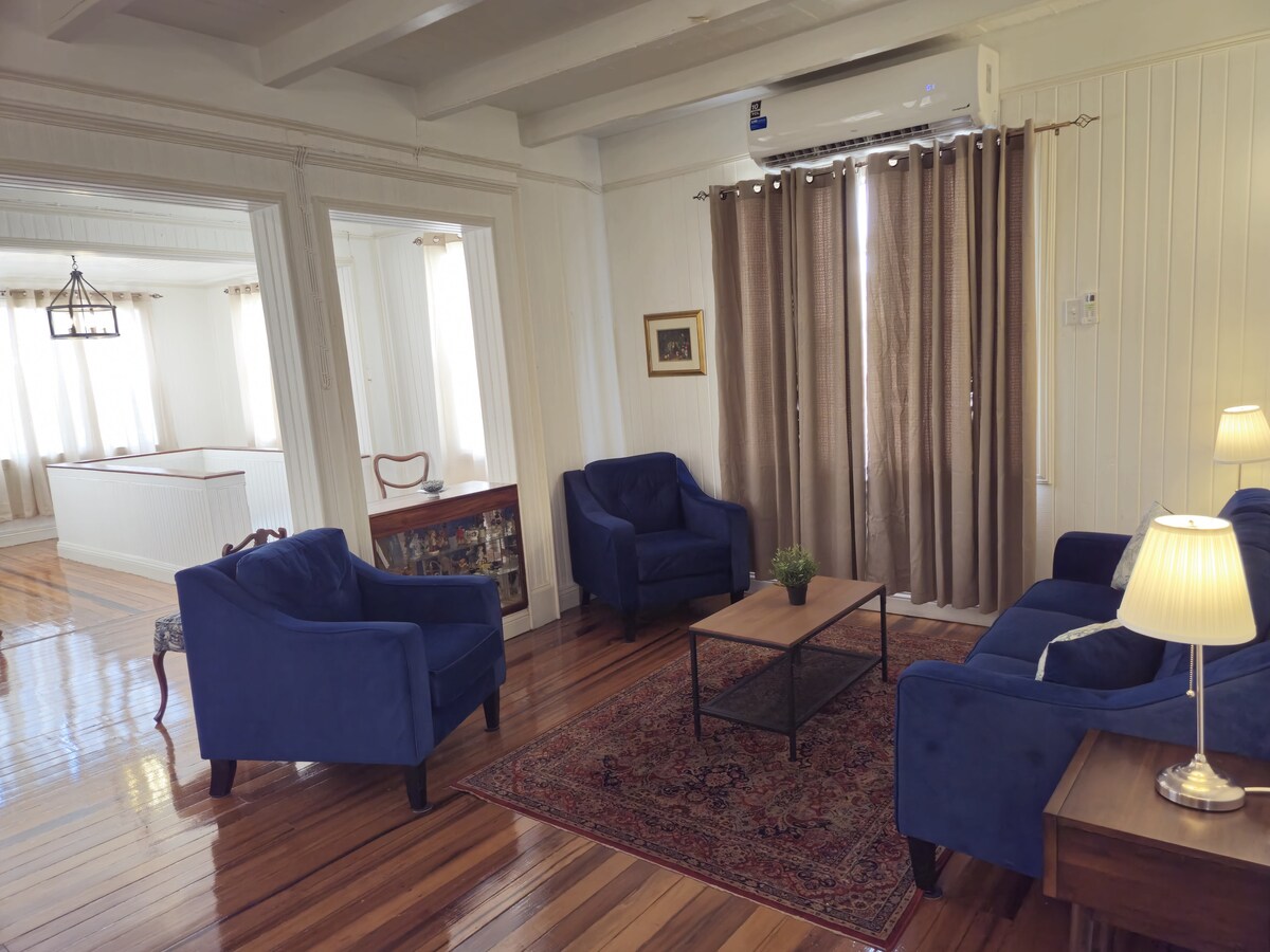 A living area features two blue armchairs facing a wooden coffee table, all set on a traditional area rug. Light-colored walls and large windows allow natural light to brighten the space, while beige curtains add a soft touch. An air conditioning unit is mounted above for comfort.