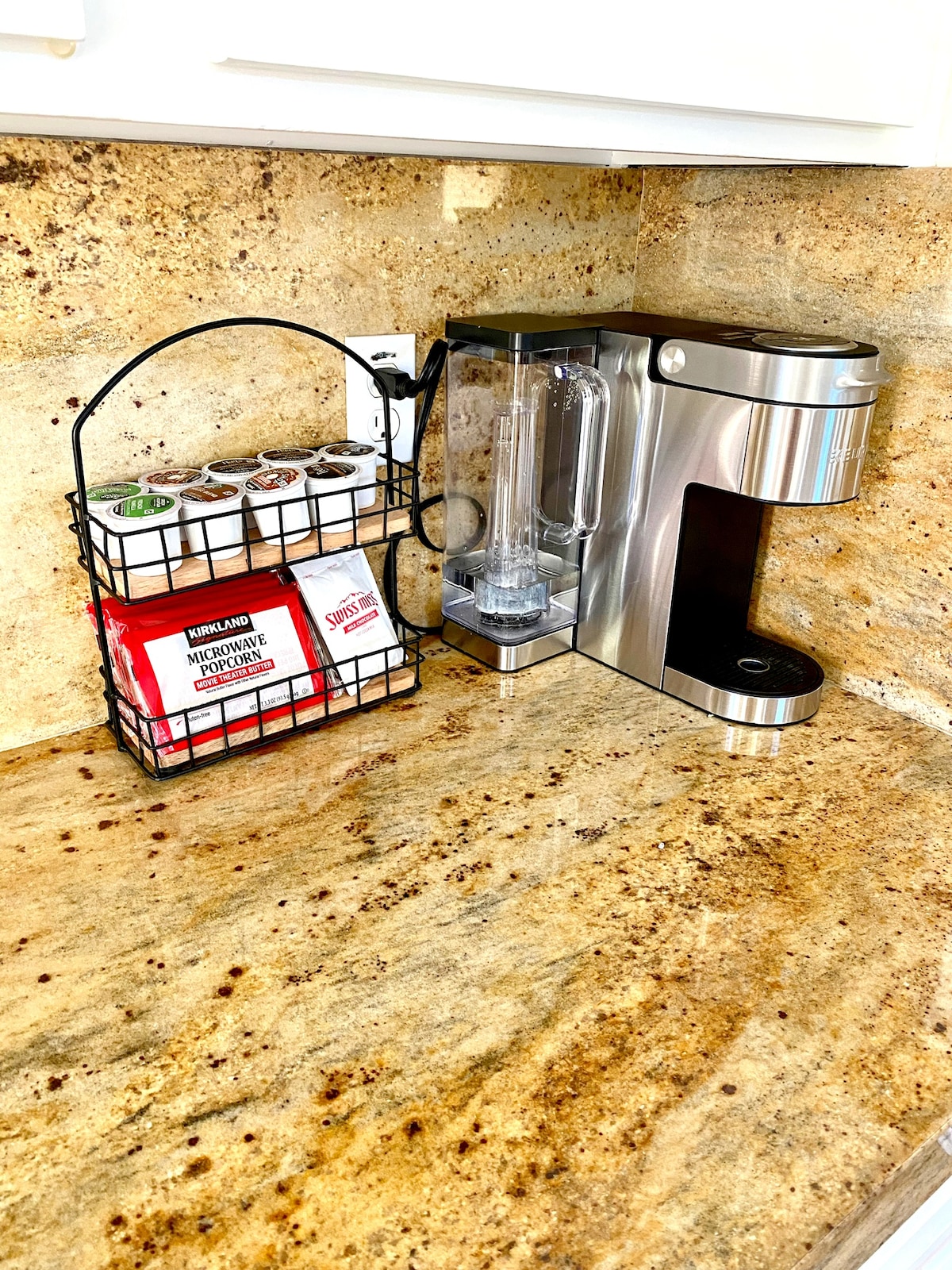 A countertop is showcased, featuring a coffee maker and a blender. To the left, a wire basket holds various single-serve coffee pods. The surface is made of a light-colored granite that highlights the appliances.