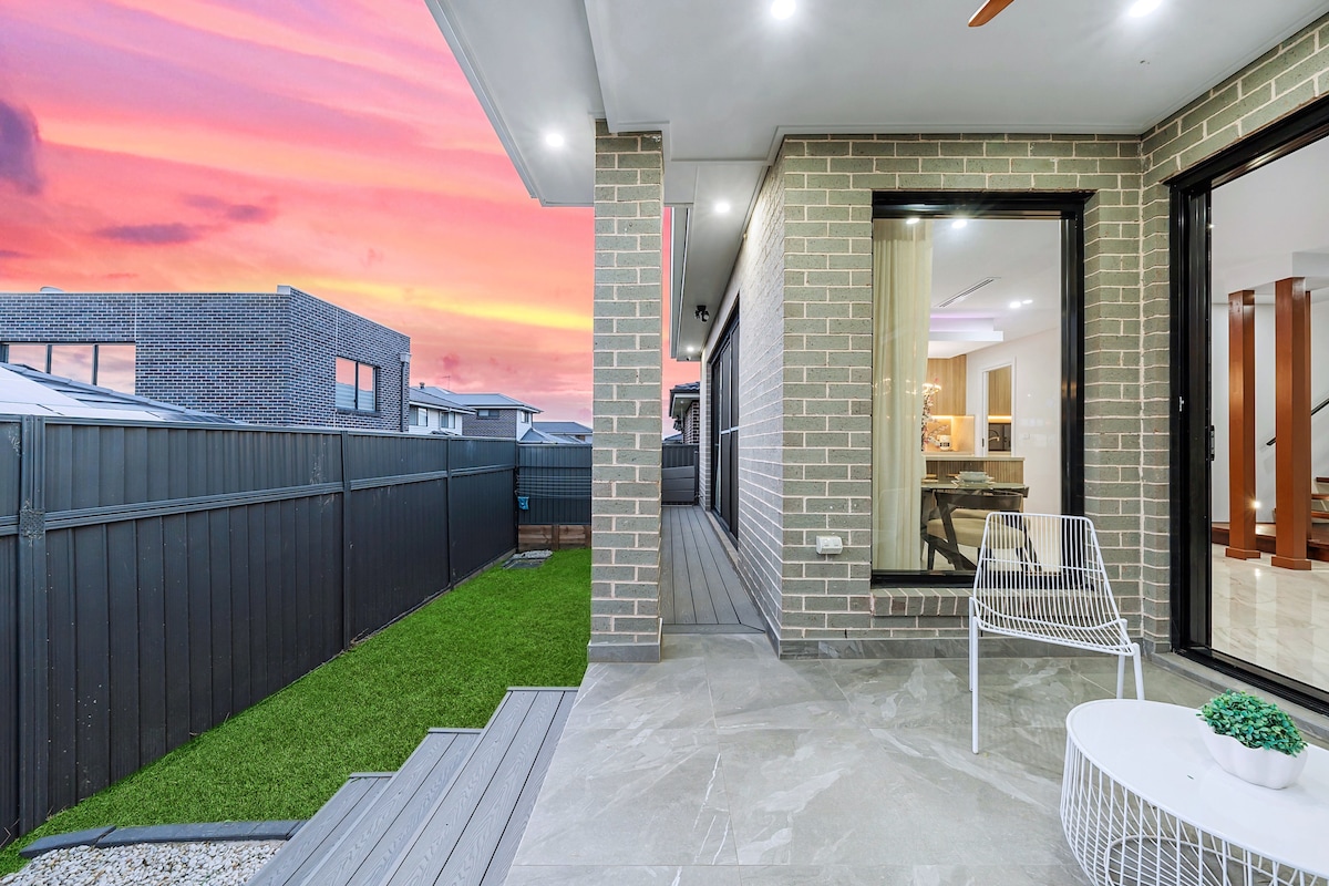 An outdoor patio area features modern grey tiles and a small white side table, accompanied by a stylish white chair. Lush green grass borders the space, and sliding glass doors reveal a warm interior. A vibrant sunset creates a colorful backdrop in the evening sky.