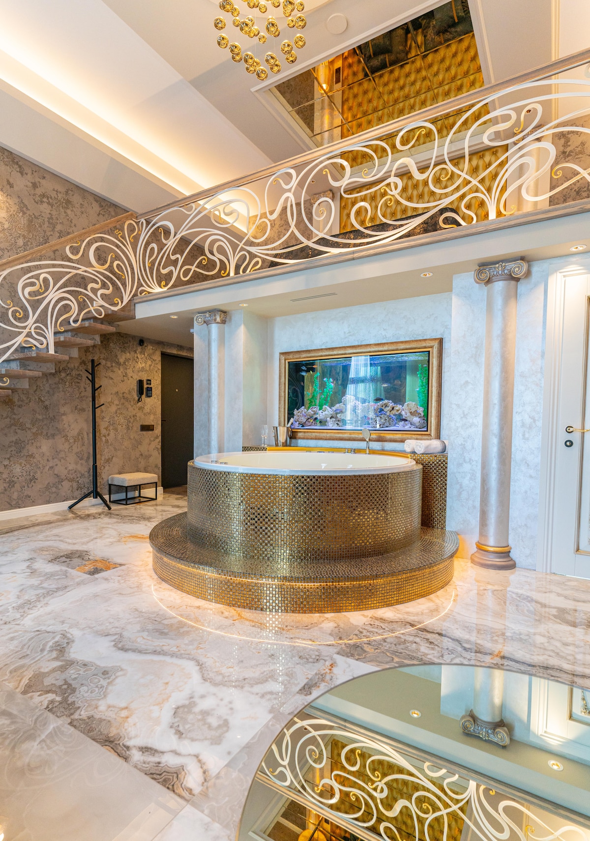 A stylishly designed area features a circular jacuzzi with a gold-toned finish, positioned on a polished marble floor. A colorful aquarium is displayed in a frame nearby, surrounded by ornate pillars and elegant wall decorations, contributing to the luxurious ambiance.