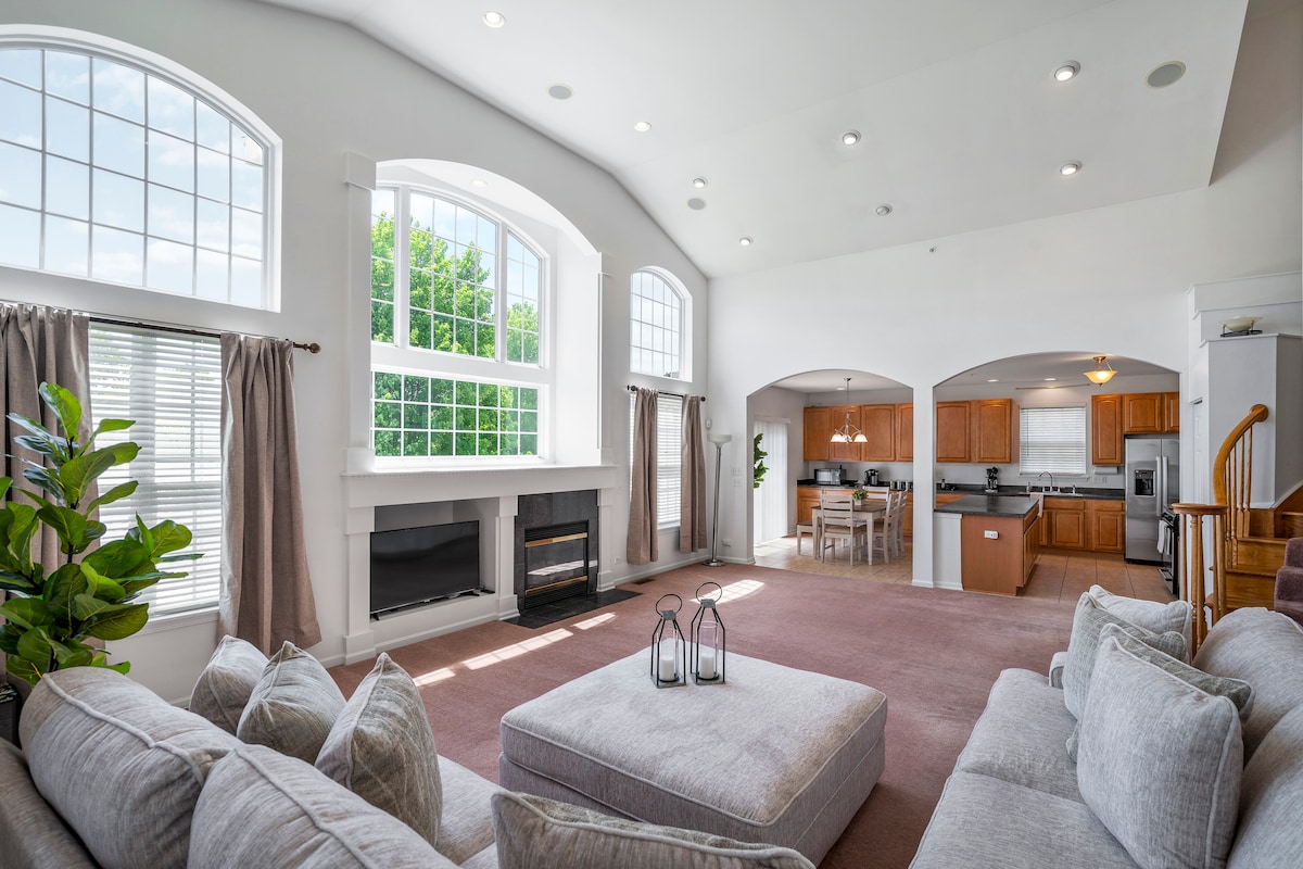 A bright living room features high ceilings and large arched windows, allowing natural light to flow in. A comfortable sectional sofa is positioned around a plush ottoman. A modern fireplace and leafy plants add to the inviting space. The open dining area and kitchen are visible in the background.
