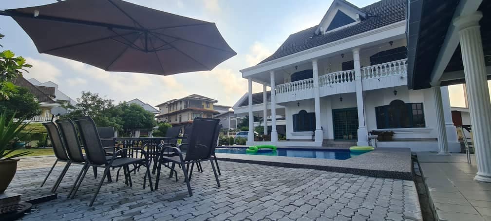 A spacious outdoor area features a swimming pool surrounded by a stone deck. Six chairs and a dining table are positioned under a large umbrella, offering shade. The villa showcases a traditional architectural style with a covered balcony and pillars.