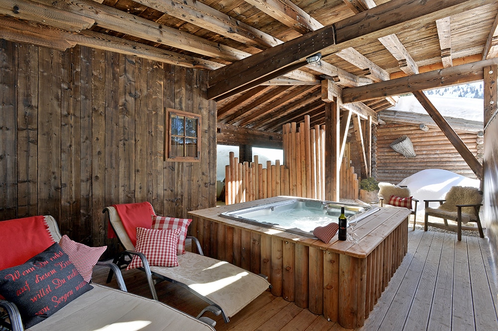The wellness area features a wooden structure with a heated outdoor whirlpool for two. Lounge chairs are present, complemented by cozy cushions. Natural light enters through windows and reflects off the wooden beams of the ceiling, creating a warm, inviting space.