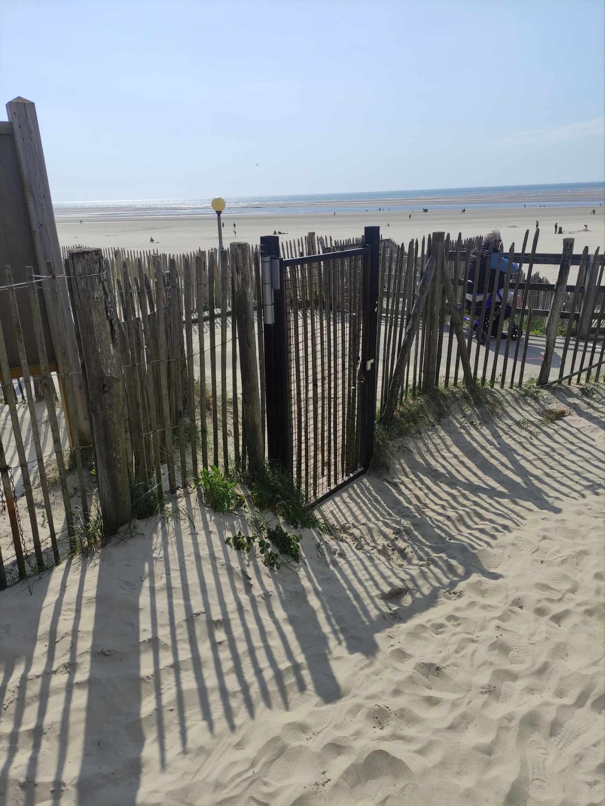 A beach entrance is framed by wooden fencing and sandy pathways leading towards the shoreline. Gentle shadows extend across the sand from the fence, suggesting a serene atmosphere. The distant water is visible, contributing to an inviting coastal view.