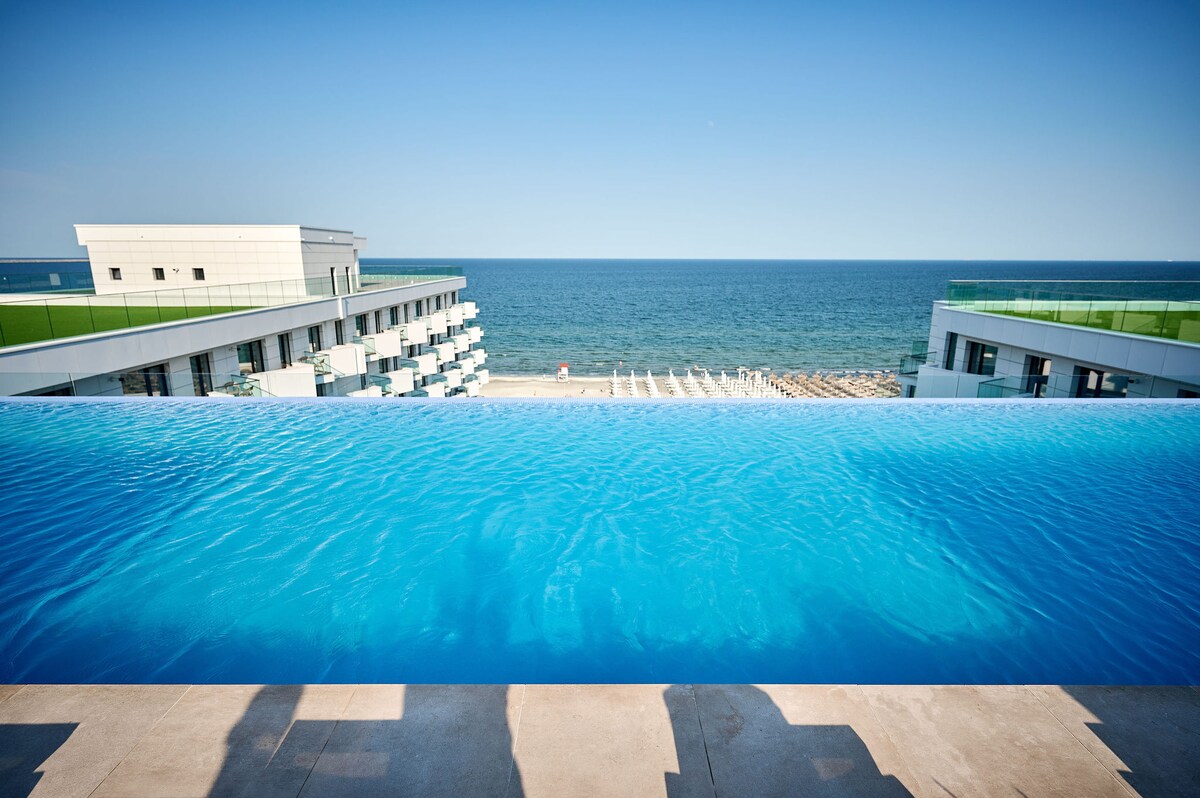 An infinity pool extends towards the horizon, merging with the serene blue ocean. Sun loungers are arranged along the beach in the distance, while the clear sky complements the tranquil atmosphere. The modern resort architecture flanks the pool, enhancing the coastal view.