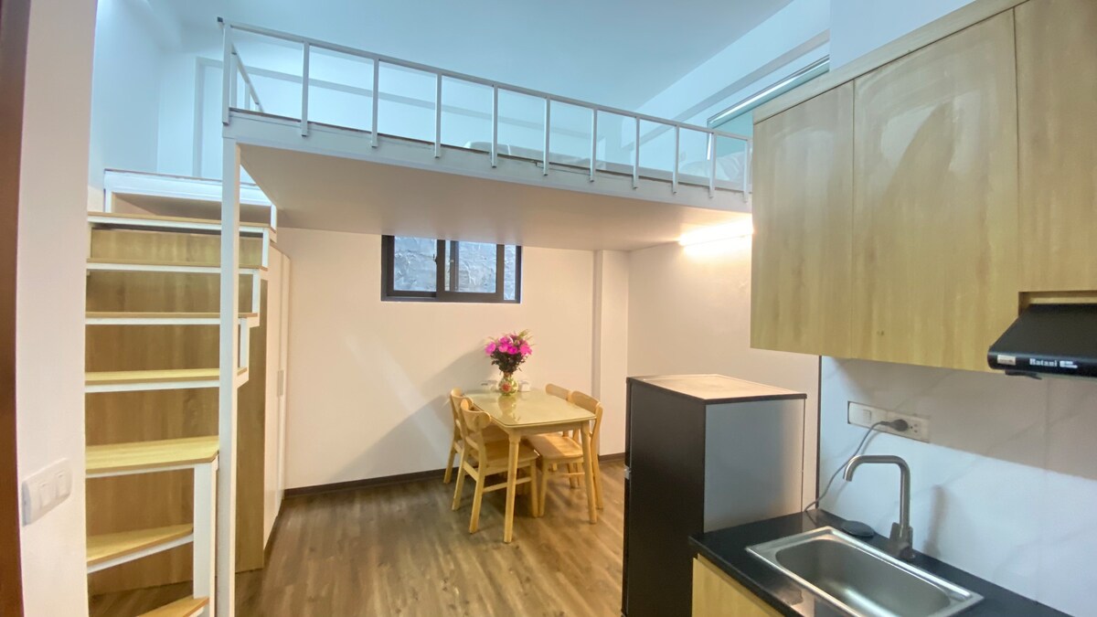 An open living space features a kitchen area with dark countertops and modern appliances. A small dining table with four wooden chairs is positioned beside a window. A wooden staircase leads to a lofted sleeping area, enhancing the room's verticality.