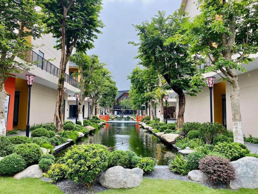 A serene courtyard is framed by lush greenery and neatly arranged bushes. A tranquil water feature runs through the center, reflecting the surrounding trees and pathways. Colorful seating areas are positioned along the edges, inviting relaxation amidst a harmonious landscape.