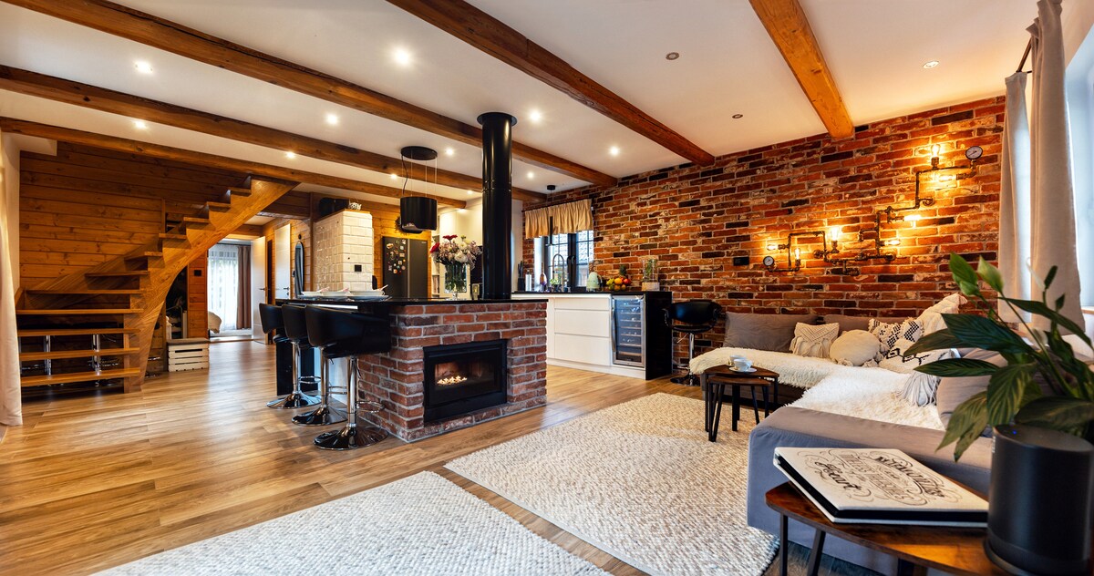 An open-plan living space features exposed brick walls and warm wooden beams. A cozy seating area is arranged near a contemporary fireplace. The well-equipped kitchen and dining area can be seen in the background, while natural light floods through large windows.