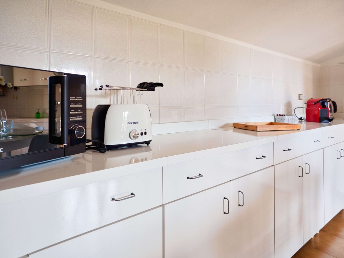 A modern kitchen is presented with sleek white cabinetry and a smooth countertop. Appliances such as a microwave, toaster, and coffee machine are visible. The backsplash features white tiles that enhance the bright and clean environment, contributing to a functional cooking space.