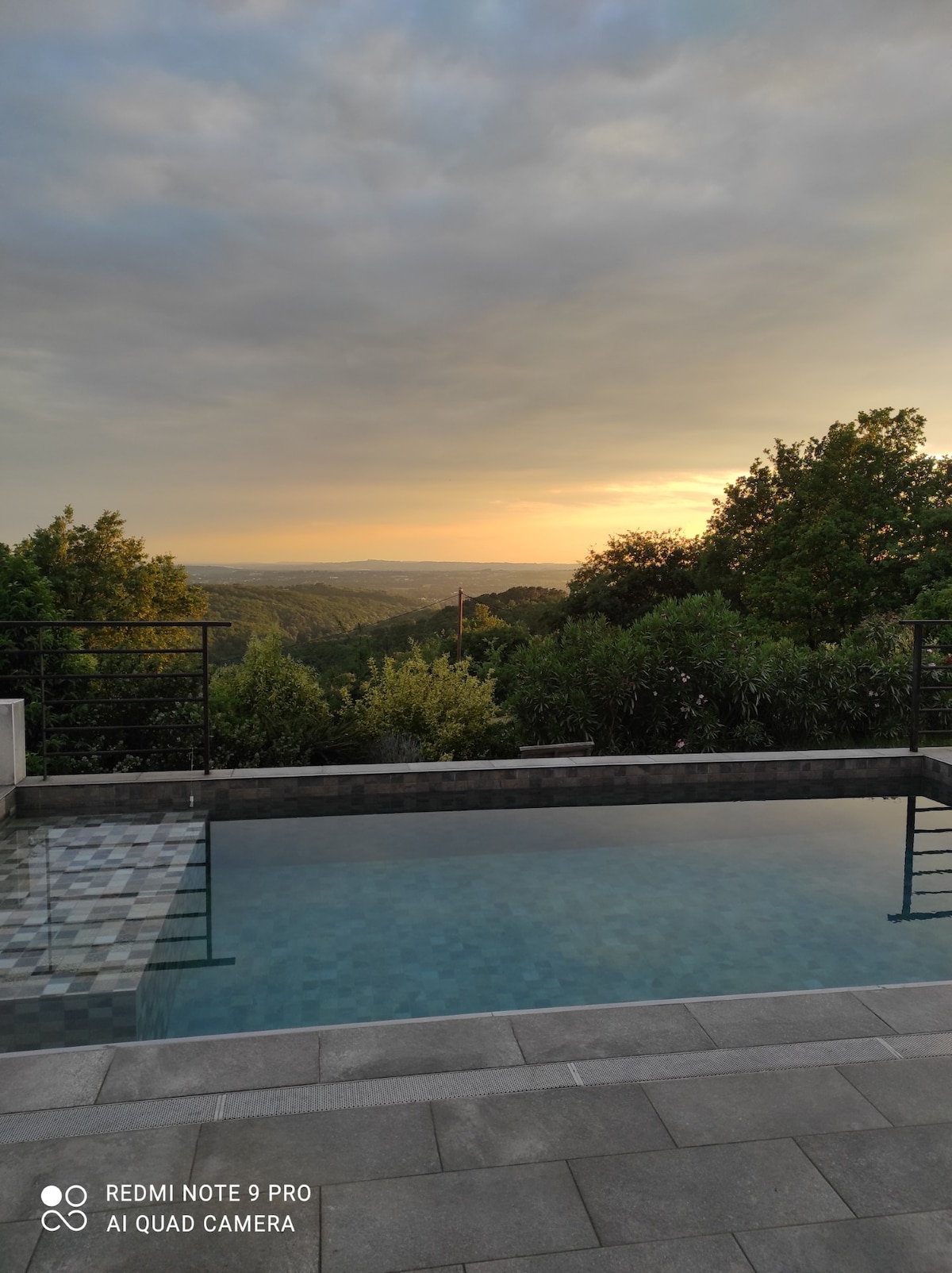 A serene infinity pool reflects the soft hues of a sunset, surrounded by lush greenery. The horizon features rolling hills under a gently clouded sky, creating a peaceful backdrop. Steps lead into the clear water, inviting relaxation and tranquility.