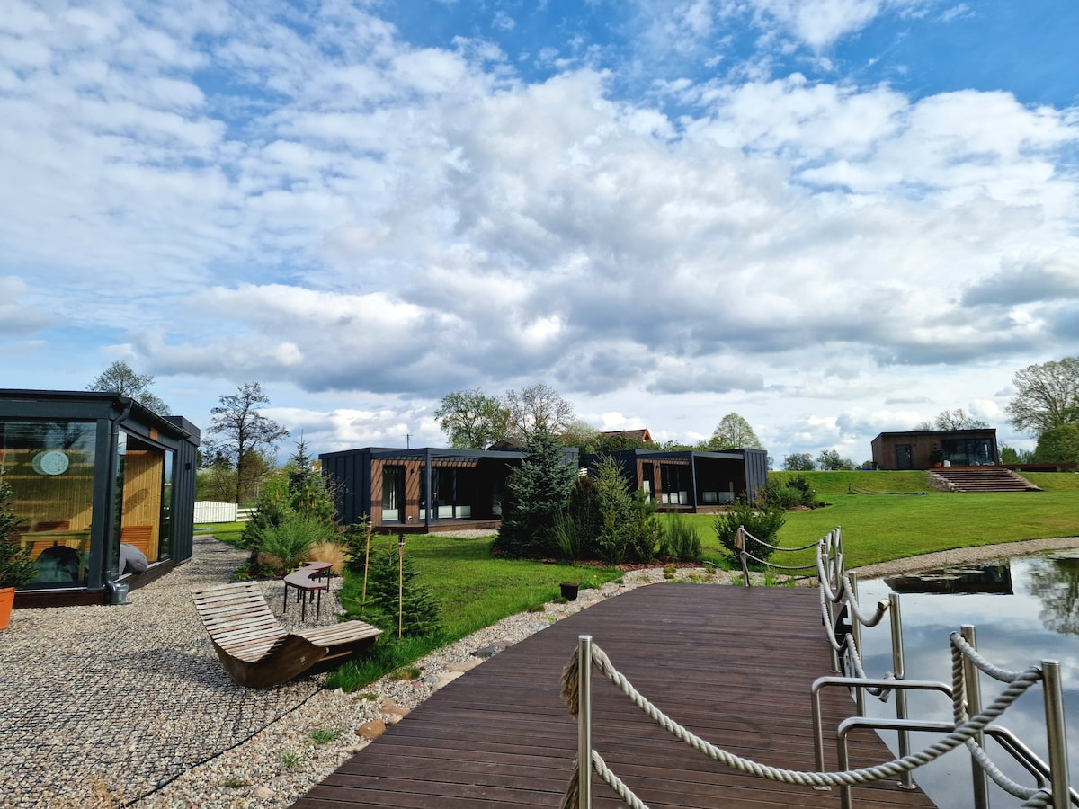 Multiple modern cabins are set against a cloudy sky, bordered by lush greenery. A wooden deck extends over a pond, featuring benches and railings along the path. The design emphasizes natural materials, creating a harmonious blend with the surrounding landscape.