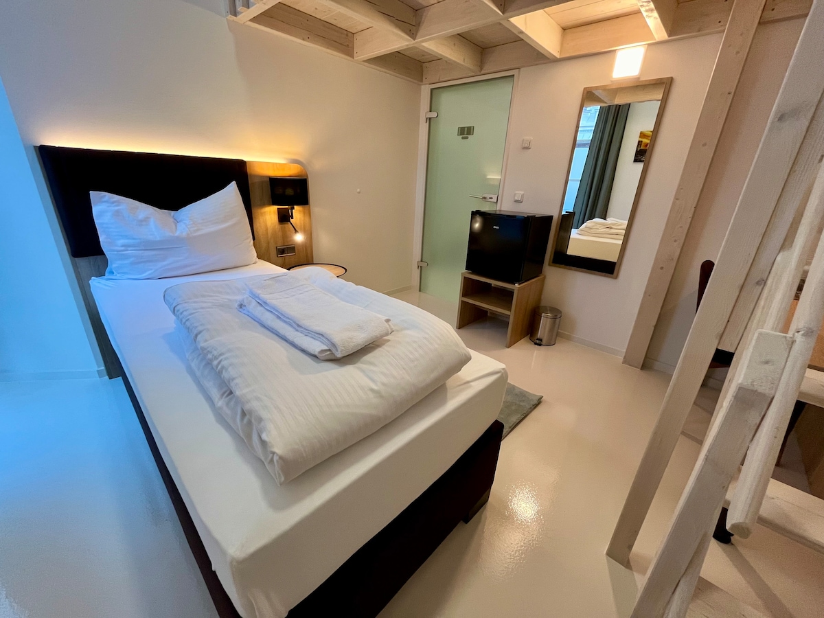 A bright sleeping area features a spacious bed with white linens and two folded towels. A flat-screen television is positioned near a mirrored dresser, and soft lighting is provided by wall-mounted lamps. A glass door leads to a partitioned area, enhancing the room's open feel.