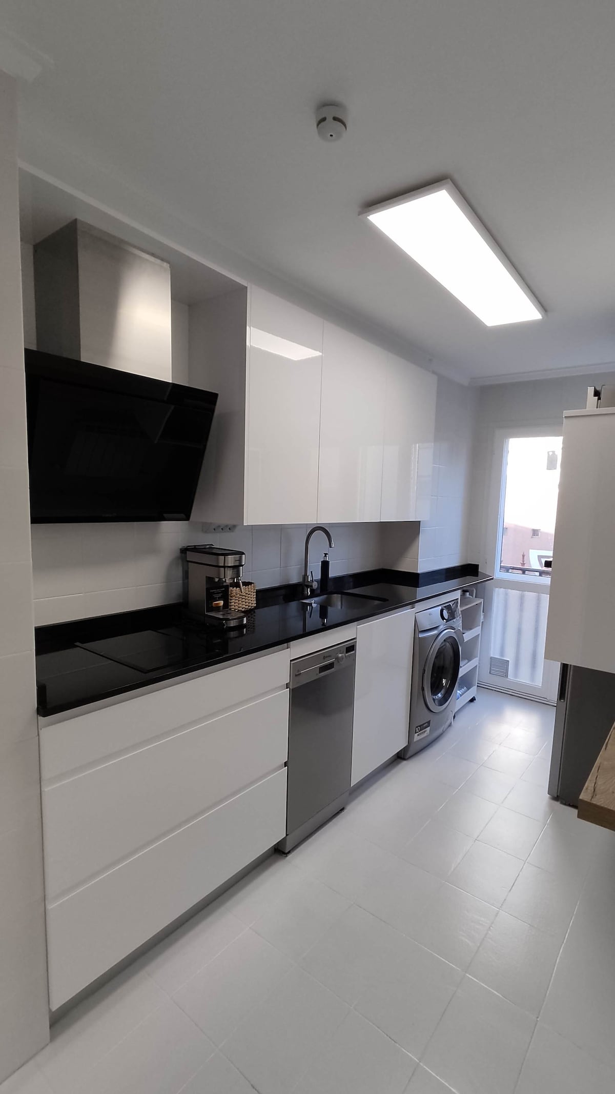 A modern kitchen features sleek white cabinetry with a contrasting black countertop. Stainless steel appliances, including a dishwasher and washing machine, are arranged for convenience. A window allows natural light, creating a bright and functional cooking space.