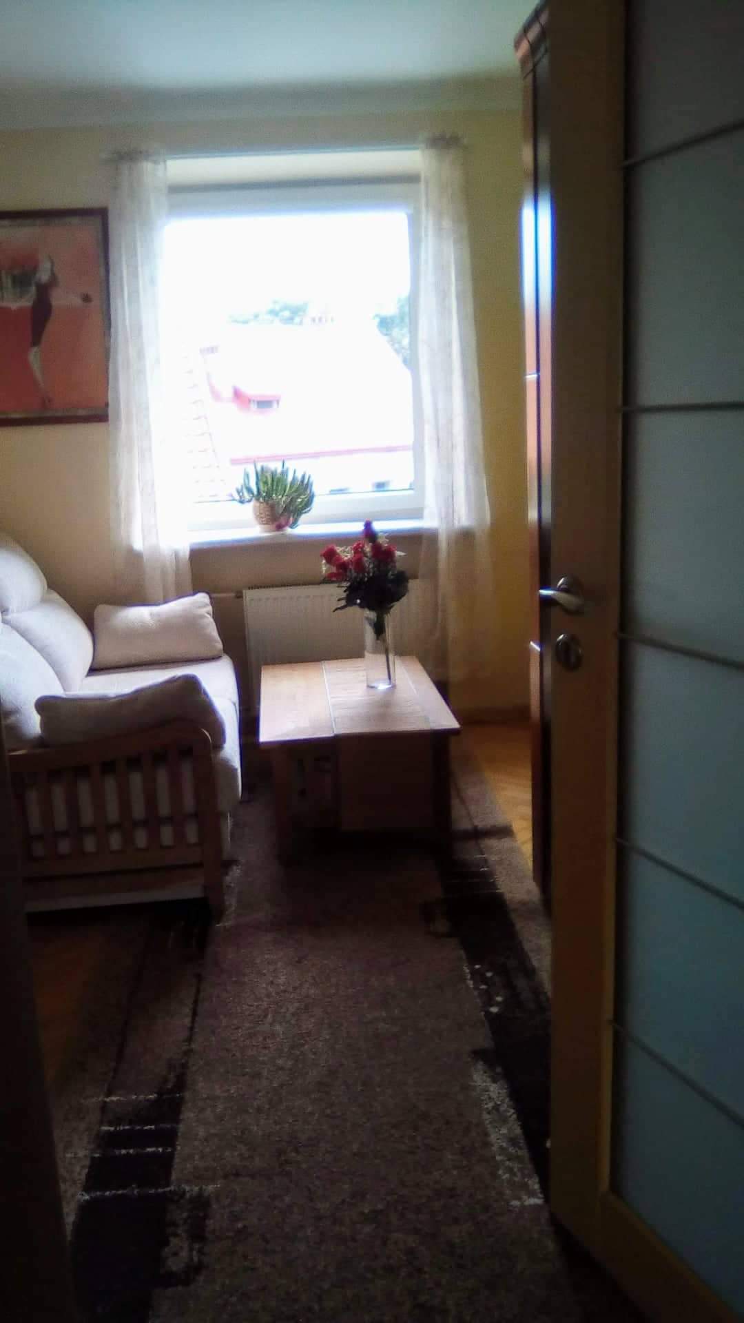 A light-filled living space is framed by a window adorned with sheer curtains. A comfortable sofa is paired with a wooden coffee table, which holds a vase of flowers. The warm wooden floors complement a subtle area rug, creating a cozy ambiance.