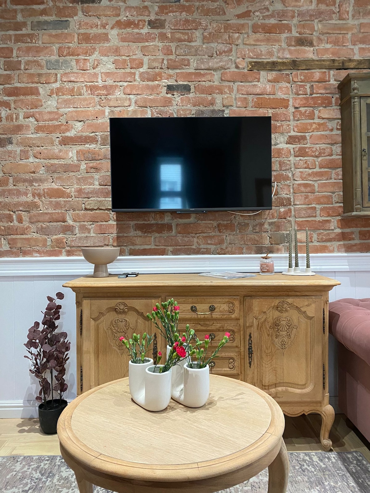 A warm living space features a wooden sideboard beneath a mounted flat-screen television. A circular wooden table is adorned with potted plants, while a brick wall adds rustic charm to the background. Decorative elements include a floor lamp and a tall cabinet in the corner.