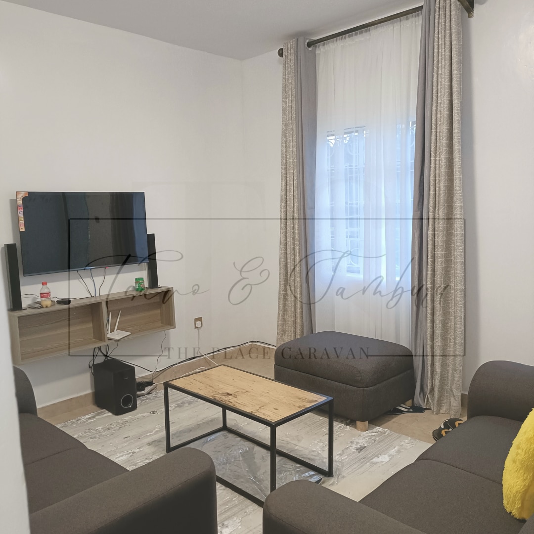 A spacious living area is shown with modern furnishings. Two comfortable sofas and a wooden coffee table are positioned around a flat-screen TV mounted on the wall. Natural light filters through sheer curtains, enhancing the welcoming atmosphere.
