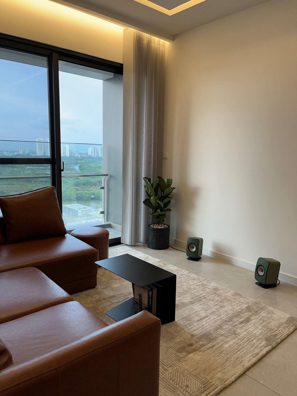 A cozy corner of the living area features a brown leather sofa paired with a sleek black side table. A tall green plant stands in a pot beside a large window, offering views of the cityscape beyond. Subtle lighting complements the warm tones of the furnishings.