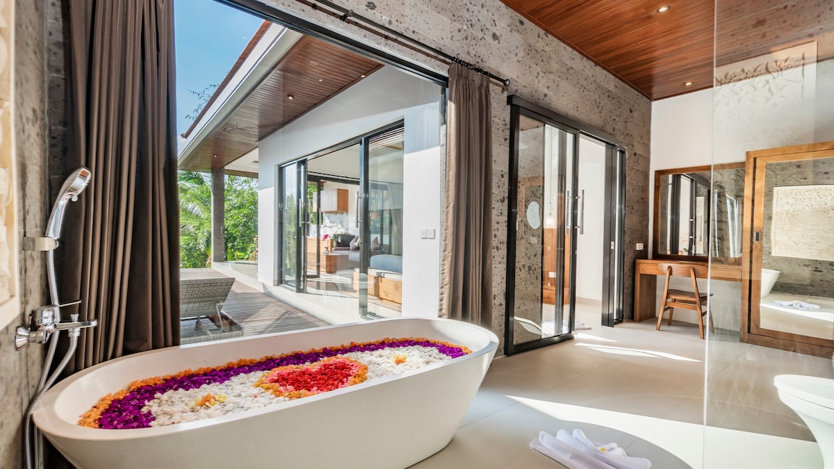 A spacious bathroom features a freestanding bathtub adorned with colorful flower petals, creating a calming focal point. Glass doors provide views of the outdoor area, blending natural light and lush greenery with the stylish interior. Soft towels are neatly placed nearby.