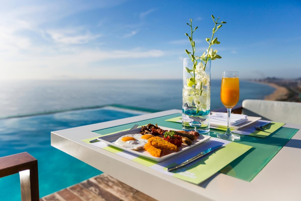 A dining table is set with a plate of breakfast items, including fried eggs and golden-brown croquettes. A glass of orange juice sits next to a flower vase filled with white orchids. The backdrop features an expansive sea view, with a rooftop infinity pool blending into the horizon.