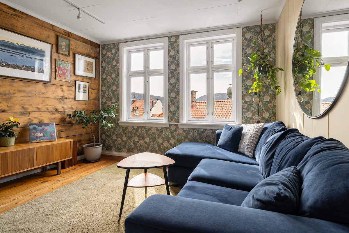 A cozy living area is highlighted by a large blue sectional sofa and a round coffee table. Bright light streams through multiple windows, revealing patterned wallpaper and wooden paneling. A vibrant plant adds a touch of greenery alongside a low wooden cabinet and framed art on the walls.