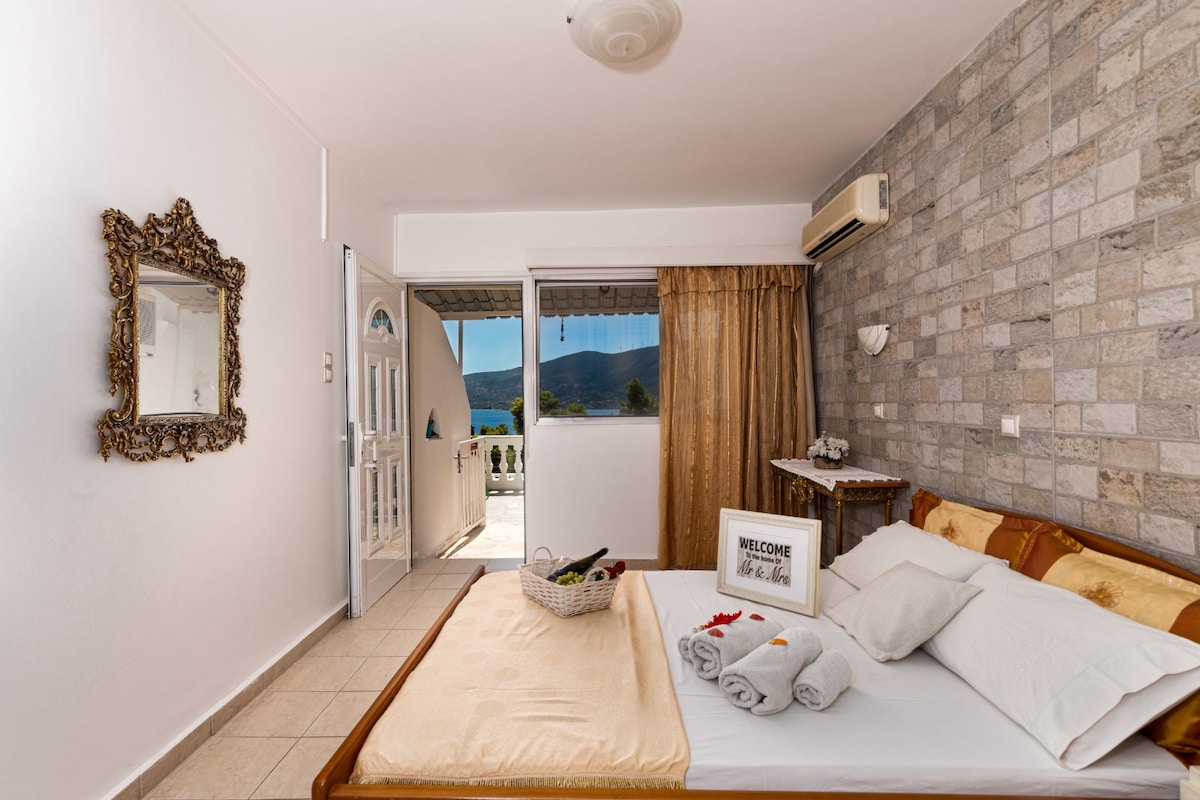 A bright bedroom features a spacious bed adorned with rolled towels and decorative rose petals. Natural light streams through the open door, revealing a scenic view. A vintage mirror and a small table with a welcome sign are positioned against a textured stone wall, adding character.