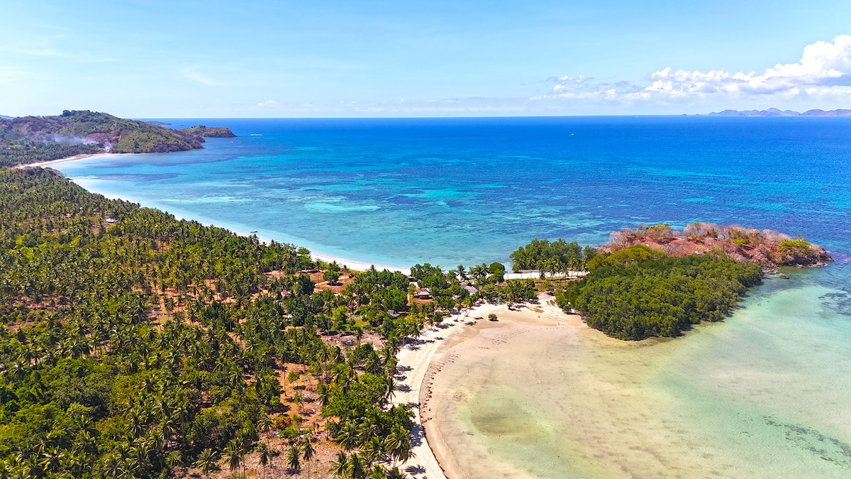 Aerial view showcases the tranquil twin beaches lined with swaying palm trees. The clear turquoise waters gently transition to deeper blue shades, contrasting with the lush greenery of the nearby hills. The serene landscape embodies a peaceful tropical getaway.